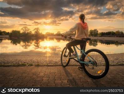 Woman riding a mountain bike near lake at sunset in summer. Colorful landscape with sporty girl riding a bicycle, beach, beautiful sky reflected in water in park in spring. Sport and travel. Biking