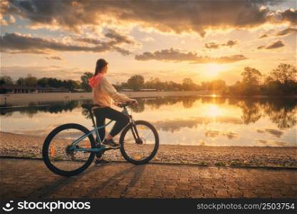 Woman riding a mountain bike near lake at sunset in summer. Colorful landscape with sporty girl riding a bicycle, beach, beautiful sky reflected in water in park in spring. Sport and travel. Biking