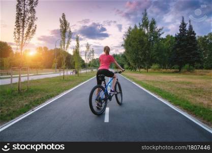 Woman riding a mountain bike on the asphalt road at sunset in summer. Colorful landscape with sporty girl riding a bicycle, road, green trees, grass and gold sunlite in park. Sport and travel. Cycle