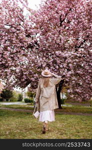 woman&rsquo;s day. Beautiful woman stands with her back near the sakura trees. Woman in hat, dress and stylish coat. Pink flowers blooming in Uzhhorod, Ukraine. Blossom around. Spring time concept. woman&rsquo;s day. Beautiful woman stands with her back near the sakura trees. Woman in hat, dress and stylish coat. Pink flowers blooming in Uzhhorod, Ukraine. Blossom around. Spring time concept.