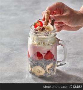 Woman&rsquo;s hand decorates healthy layered dessert with yogurt, chia pudding, banana and strawberry in glass on a stone background. Layered berry and chia seeds smoothies in a glass jar, girls hand decorate cookies on a gray concrete background.
