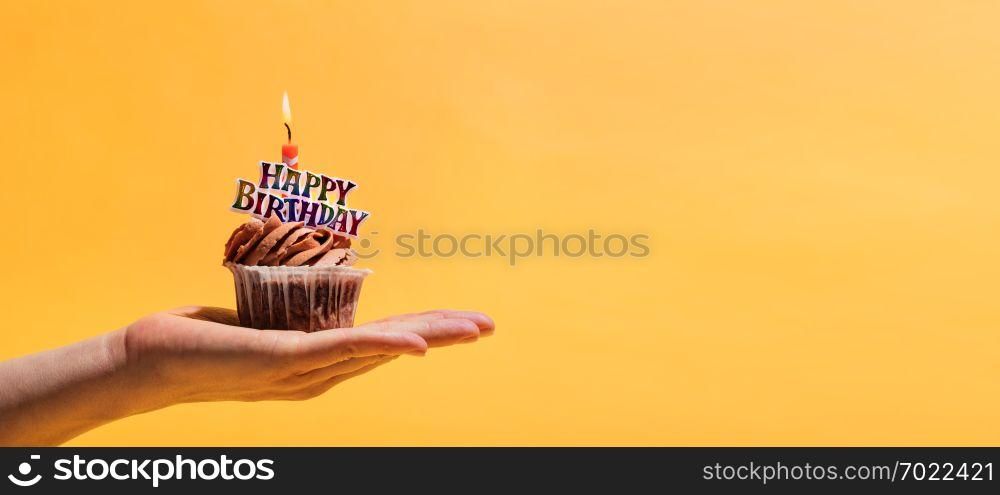 Woman&rsquo;s hand holding decorated birthday muffin against yellow background. Birthday party and celebration.. Woman&rsquo;s hand holding decorated birthday muffin.