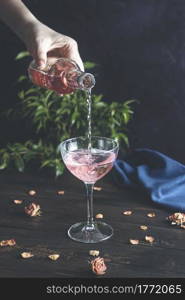 Woman&rsquo;s hand holds a bottle and pouring champagne in elegant wineglass on dark wooden table surface with dry roses, close up, shallow depth of the field