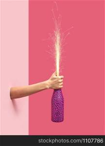 Woman&rsquo;s hand holds painted champagne bottle with holiday firecracker on a duotone pink background, copy spase. Christmas concept.. Holiday firework in a painted bottle in a female&rsquo;s hand.