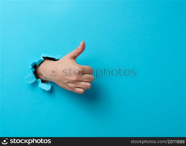 woman&rsquo;s hand sticks out of the torn hole and shows a gesture okay, thumb up. Blue background