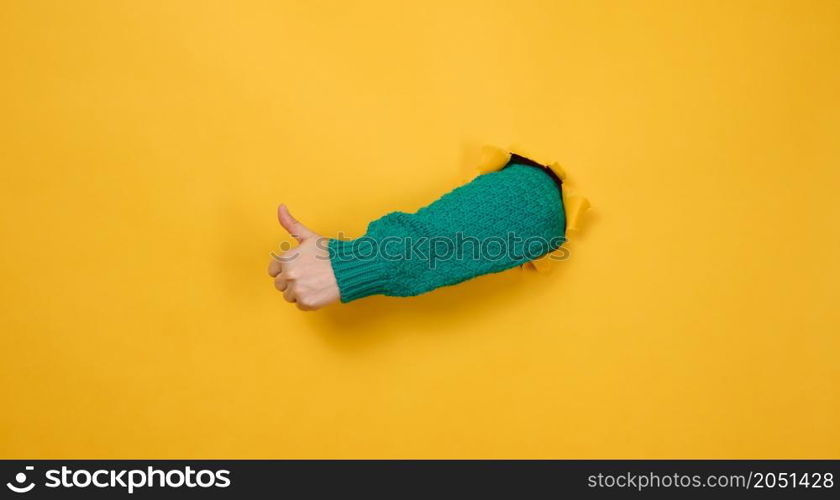 woman&rsquo;s hand sticks out of the torn hole and shows a gesture okay, thumb up. Yellow background