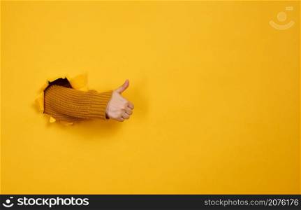 woman&rsquo;s hand sticks out of the torn hole and shows a gesture okay, thumb up. Yellow background, copy space