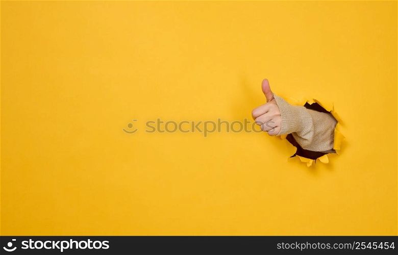woman&rsquo;s hand sticks out of the torn hole and shows a gesture okay, thumb up. Yellow background