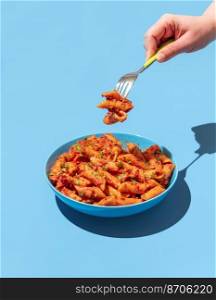 Woman&rsquo;s hand taking pasta with a fork from a blue bowl, minimalist on a blue table. Pasta alla arrabbiata in a blue bowl in bright light.