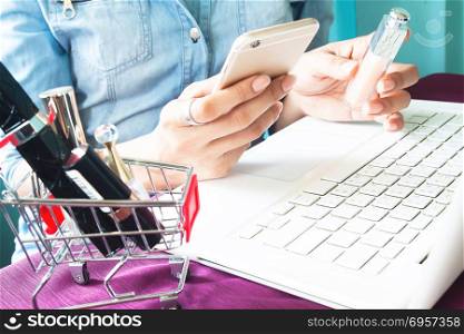 Woman&rsquo;s hand using smartphone and holding lipstick with beauty i. Woman&rsquo;s hand using smartphone and holding lipstick with beauty items in shopping cart, Online shopping