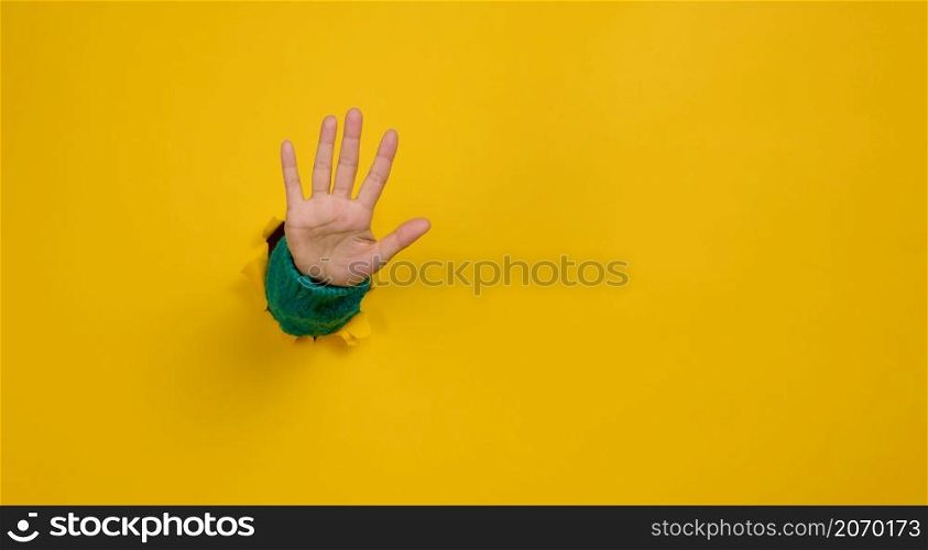 woman&rsquo;s hand with an open palm sticks out of a torn hole in yellow paper, gesturing to stop. Copy space
