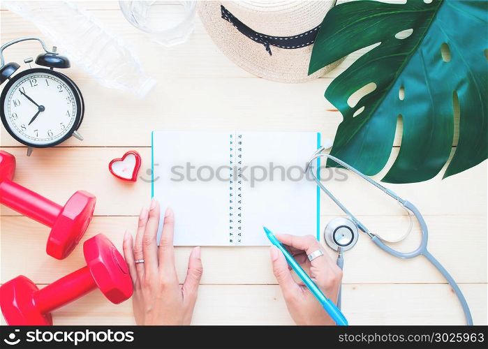 Woman&rsquo;s hand writing on blank notebook with sport and medical equipments on wooden table, Woman&rsquo;s health and lifestyle concept