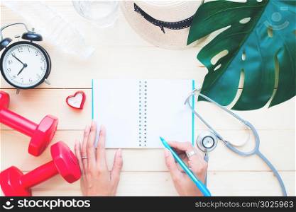 Woman&rsquo;s hand writing on blank notebook with sport and medical equipments on wooden table, Woman&rsquo;s health and lifestyle concept