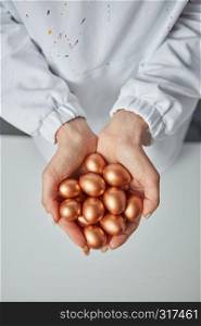 Woman's hands hold small painted golden eggs around a gray background. Wealth concept. Top view. Golden painted small eggs hold the hands of a woman on a gray background. Concept of profit and wealth. Top view