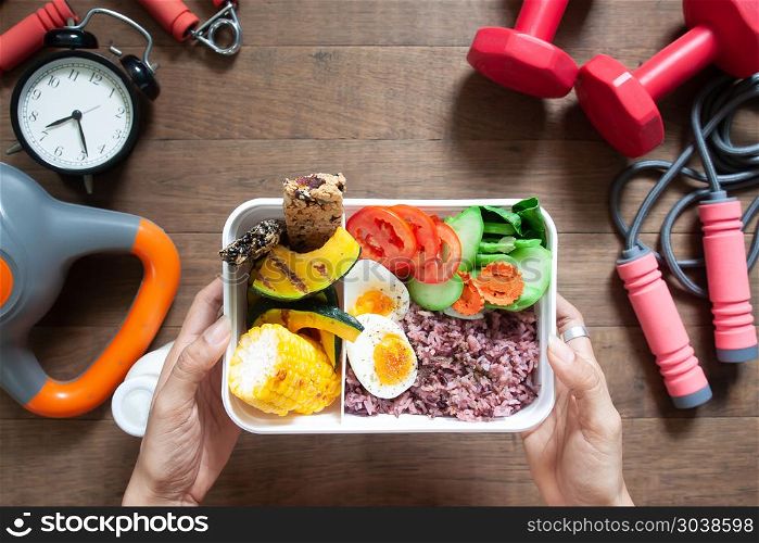 Woman&rsquo;s hands holding lunch box with rice berry, boiled eggs, sw. Woman&rsquo;s hands holding lunch box with rice berry, boiled eggs, sweetcorn, pumpkin, tomatoes and cereal bars, Top view with sport and fitness equipments on wooden background
