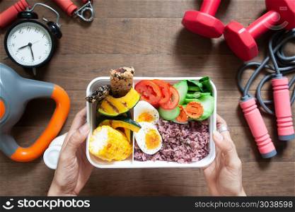Woman&rsquo;s hands holding lunch box with rice berry, boiled eggs, sw. Woman&rsquo;s hands holding lunch box with rice berry, boiled eggs, sweetcorn, pumpkin, tomatoes and cereal bars, Top view with sport and fitness equipments on wooden background