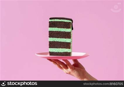 Woman’s hand holding a plate with a slice of peppermint cake against a purple background. Homemade layer cake with mint-flavored cream and chocolate topping