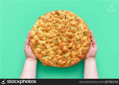 Woman’s hands hold a freshly baked rosemary focaccia above a green table. Top view with homemade focaccia with a delicious crust.
