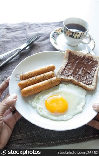 Woman's hands holding plate of breakfast with egg, sausages, bread and chocolate cream on top