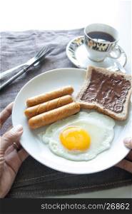 Woman's hands holding plate of breakfast with egg, sausages, bread and chocolate cream on top