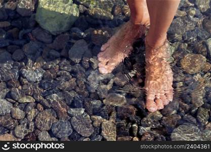 Woman?s legs ankle-deep in clear water. Ripple patterns reflect light on water over pebbles
