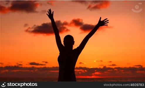 Woman's silhouette in sunset light, happy girl with raised up hands standing on the beach and enjoying warm summer evening, victory concept