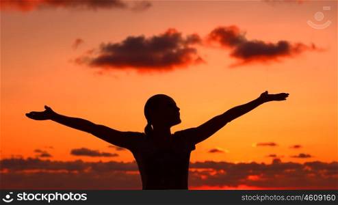 Woman's silhouette on sunset light, happy girl with raised up hands standing on the beach and enjoying warm summer evening, doing yoga exercise outdoors