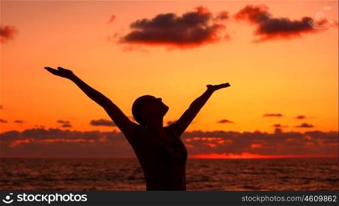 Woman's silhouette over sunset background, happy girl with raised up hands standing on the beach and looking up in the sky, enjoying summer vacation