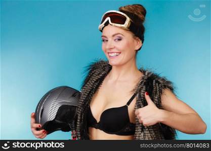 Woman seductive girl in ski googles holds helmet. Woman sexy hot skier girl wearing black bra fur vest ski googles holding helmet showing thumb up. Winter sport activity. Beautiful seductive sportswoman on blue studio shot