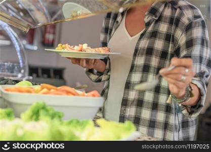 Woman serves herself food at a cafeteria holding a dish with food on her right hand and a spoon on her left hand with an out of focus background and foreground. Healthy food concept.. Woman holding a dish and serving herself