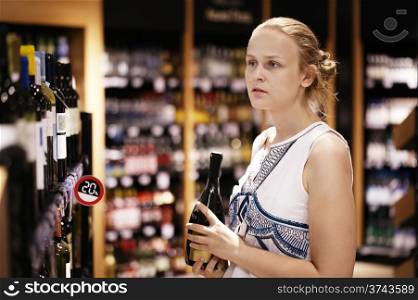 Woman shopping for wine or other alcohol in a bottle store standing in front of shelves full of bottles with a serious expression as she tries to make up her mind