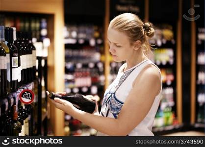 Woman shopping for wine or other alcohol in a bottle store standing in front of shelves full of bottles and holding bottle in her hand and reading inscription