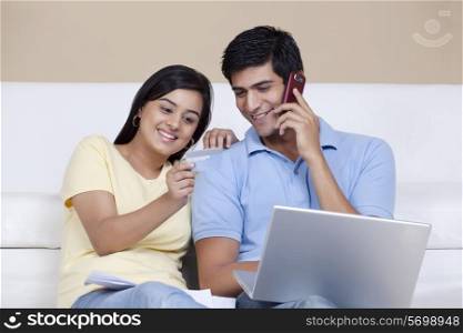 Woman showing credit card to man while talking on phone and using laptop