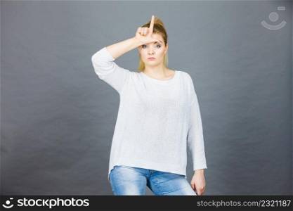 Woman showing mean sign, lame or loser gesture with L fingers on forehead, grey background.. Woman showing loser gesture with L on forehead