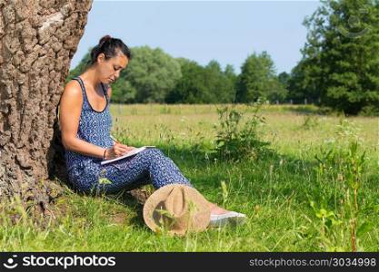 Woman sits against tree writing in meadow. Young indian woman sitting against tree trunk writing in pasture