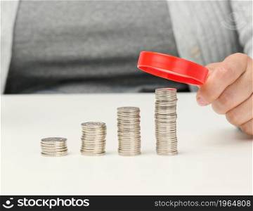 woman sits at a table and holds a red magnifying glass over stacks of white coins. The concept of budget planning, savings. Investment search