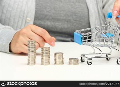woman sits at the table and holds a miniature cart and stacks of white coins. Concept of savings, sale, tax