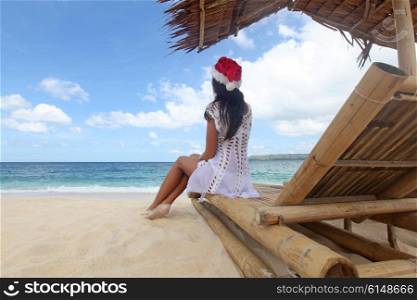 Woman sitting at chaise lounge . Woman in Santa Claus hat sitting at chaise lounge with straw parasol on white sandy beach at Philippines