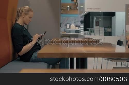 Woman sitting at the table in cafe and passing time with surfing internet on cellphone