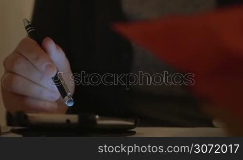 Woman sitting at the table with glass of wine in one hand. She&acute;s holding a stylus in another hand and scrolling info on the screen of her smartphone.