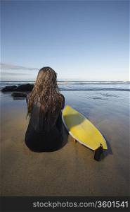 Woman sitting by surfboard on beach, back view