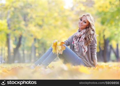 Woman sitting in autumn park. Beautiful young woman sitting and holding bunch of fall leaves in autumn park