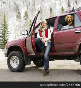 Woman sitting in dirt splattered SUV with door open as dog in back seat looks out open window in snowy countryside.