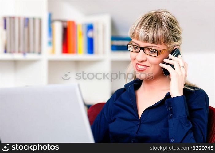 Woman sitting in front of a bookshelf using her telephone, working with a laptop in the internet from home, she is a telecommuter