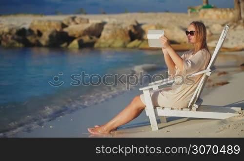Woman sitting in wooden chaise longue by the sea and taking pictures using tablet computer