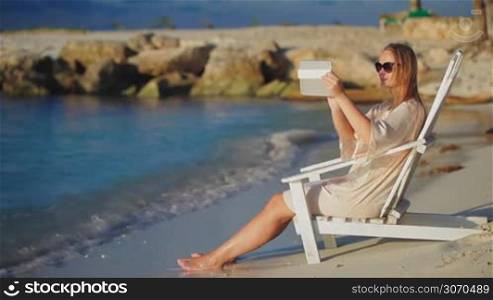 Woman sitting in wooden chaise longue by the sea and taking pictures using tablet computer
