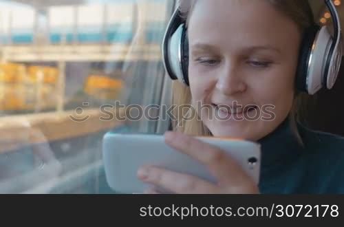 Woman sitting near the window in train and having good time. She listening to music in headphones and using smartphone