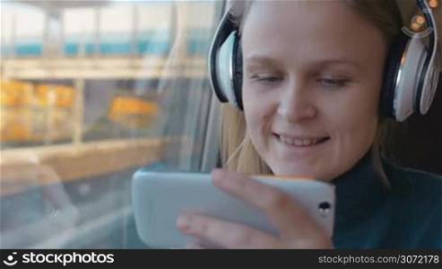 Woman sitting near the window in train and having good time. She listening to music in headphones and using smartphone