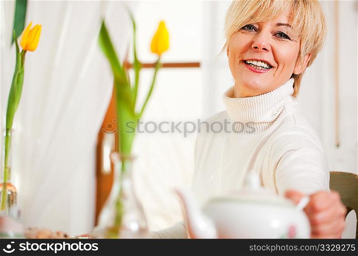 Woman sitting on a table for coffee or tea time grabbing for the coffee or tea pot, there are yellow tulip flower on the table, in the background is a window, whole scene is sunlit