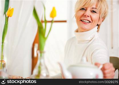 Woman sitting on a table for coffee or tea time grabbing for the coffee or tea pot, there are yellow tulip flower on the table, in the background is a window, whole scene is sunlit